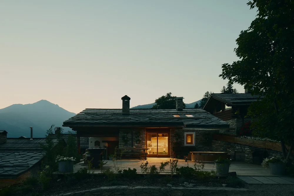 Chalet in pietra illuminato al crepuscolo, con terrazza e montagne sullo sfondo